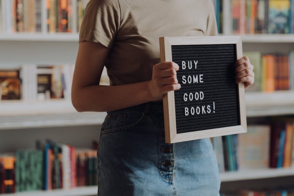 A woman holding a sign that says "buy some good books" in front of a shelf of books from a publishing imprint - Photo by cottonbro studio for pexels