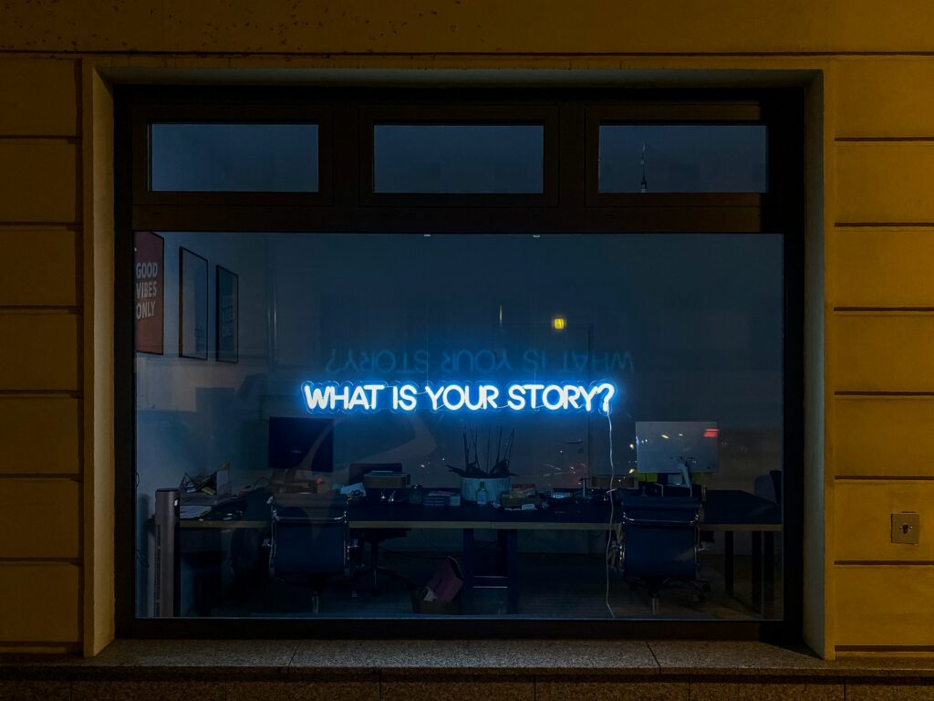 A neon signing reading What is Your Story? in a shot window - writing original fiction - Photo by Etienne Girardet on Unsplash